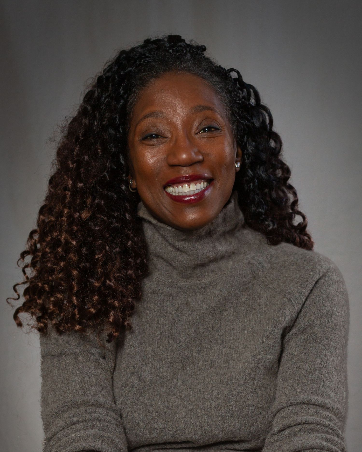A smiling woman with curly dark hair wearing a gray turtleneck poses for a professional headshot.