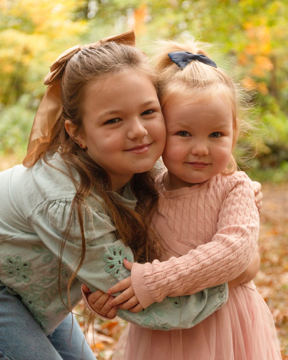 Two smiling young sisters embrace outdoors among colorful autumn trees, one wearing a hair bow and the other a pink sweater dress.