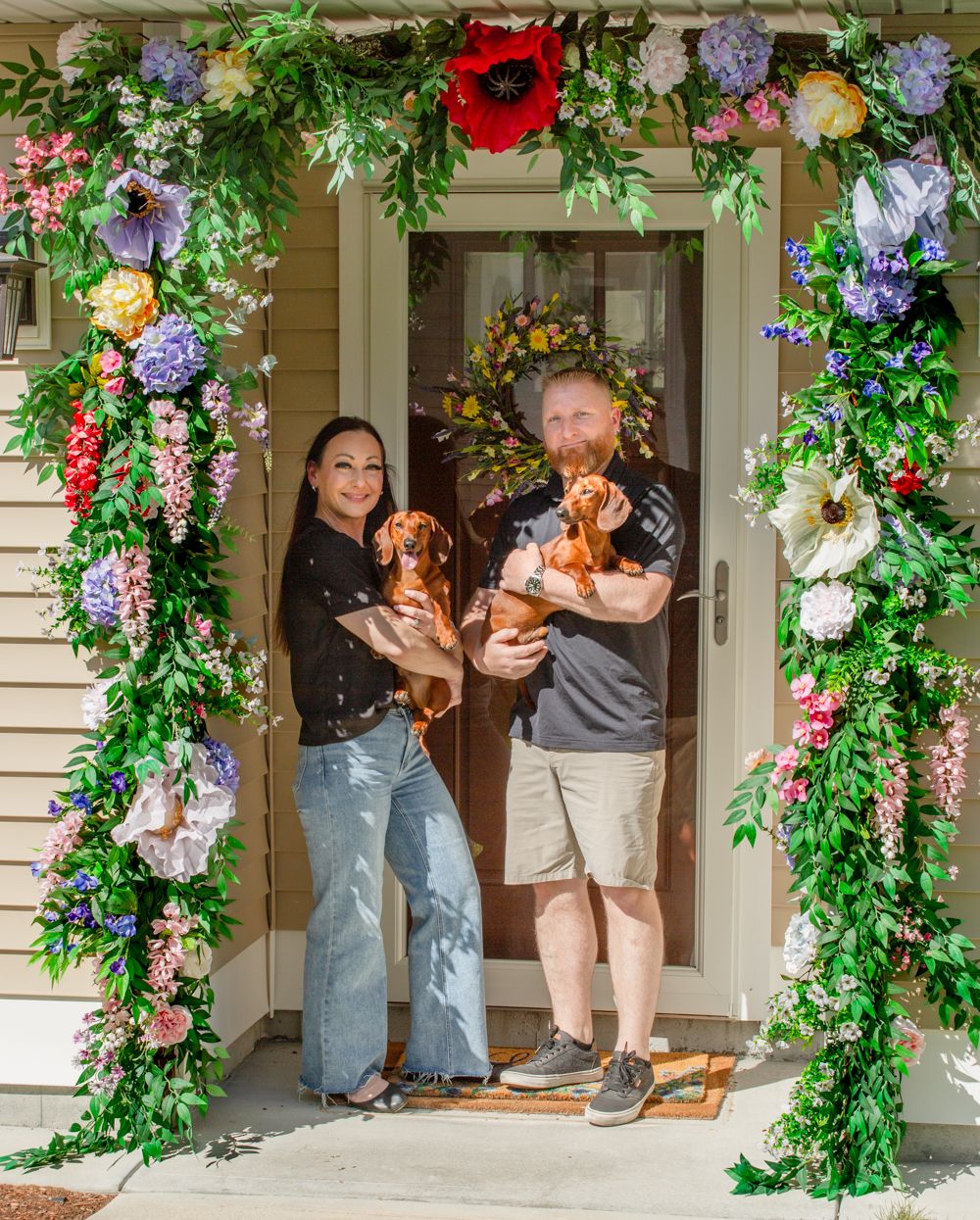 Couple holding dachshund dogs standing under a colorful floral arch at their home entrance.