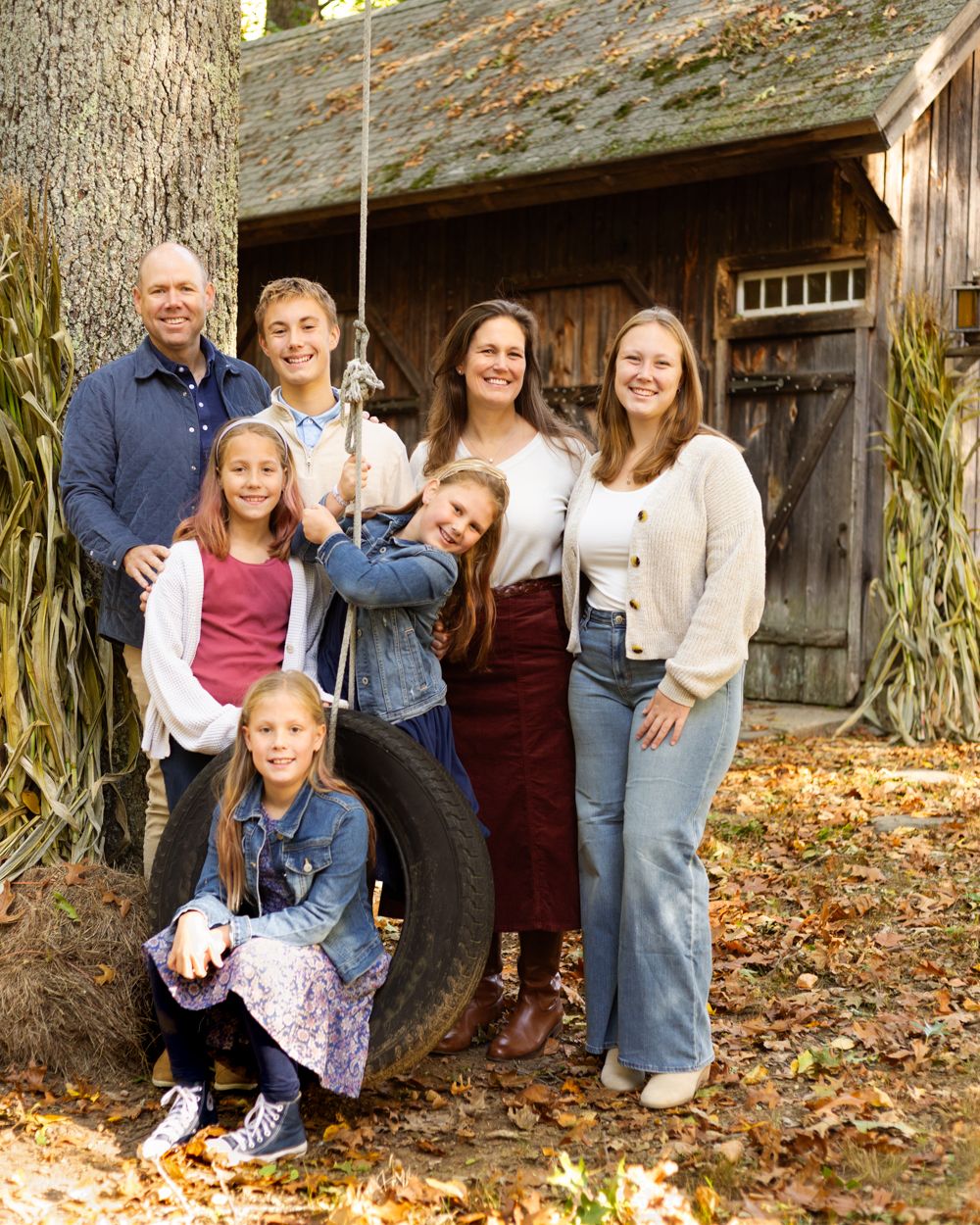 Family of six smiling together outdoors by a rustic wooden barn, posing around a tire swing among autumn leaves and cornstalks.