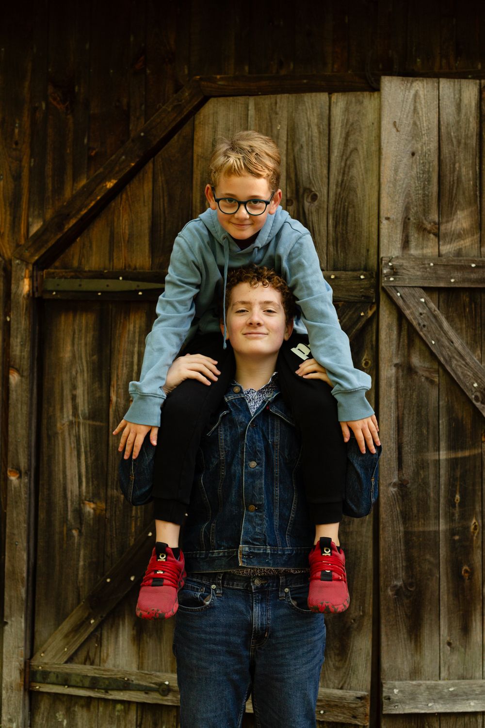 A boy in glasses and a blue hoodie sits on the shoulders of a smiling teen in a denim jacket in front of a rustic wooden barn door.