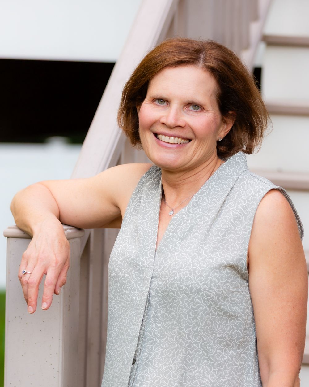 Smiling woman in sleeveless gray top leaning on white stair railing outdoors.