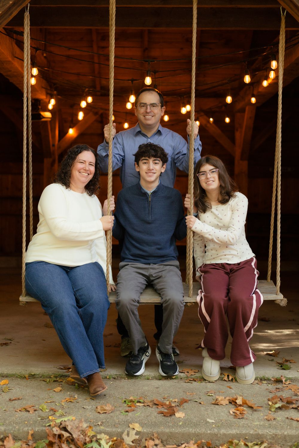 A family of four sits together on a rustic wooden swing in a barn with string lights, with fallen autumn leaves on the ground.