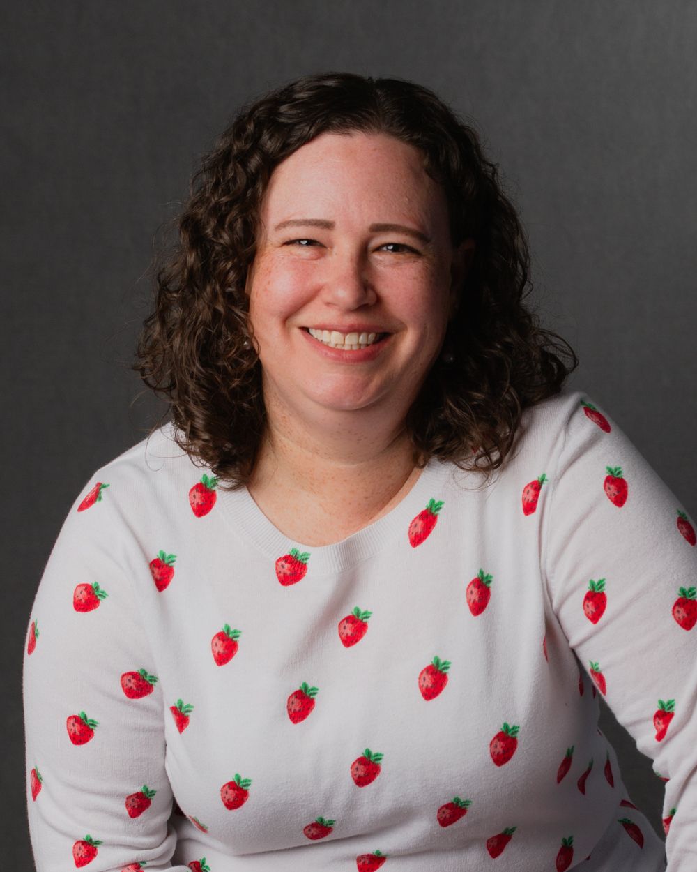Smiling woman in strawberry print top posing for professional headshot on gray background.