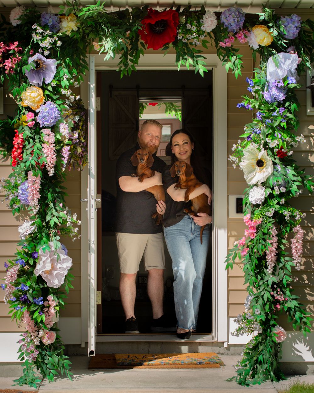 Couple holding two dachshunds standing in a doorway framed by a colorful floral arch with greenery.