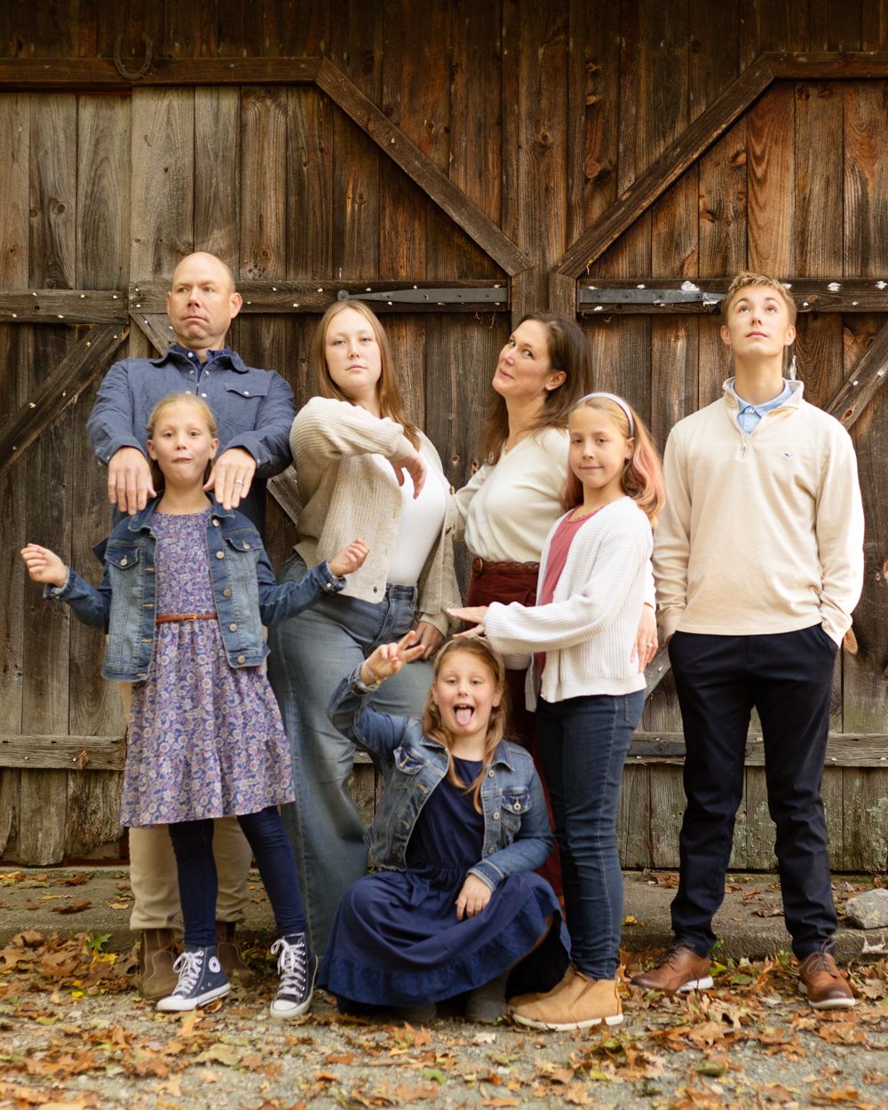 A family of six poses playfully in front of a rustic weathered barn door, with the girls making silly faces on a leaf-strewn ground.
