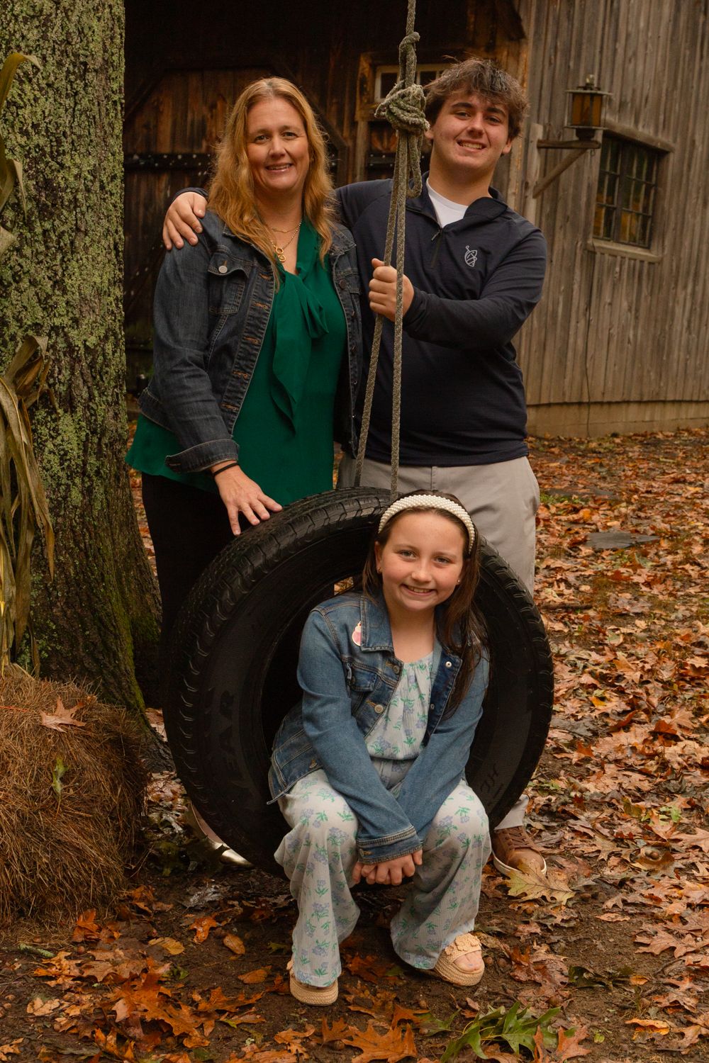 A smiling woman and teenage boy stand behind a girl in a denim jacket sitting in a tire swing surrounded by autumn leaves.