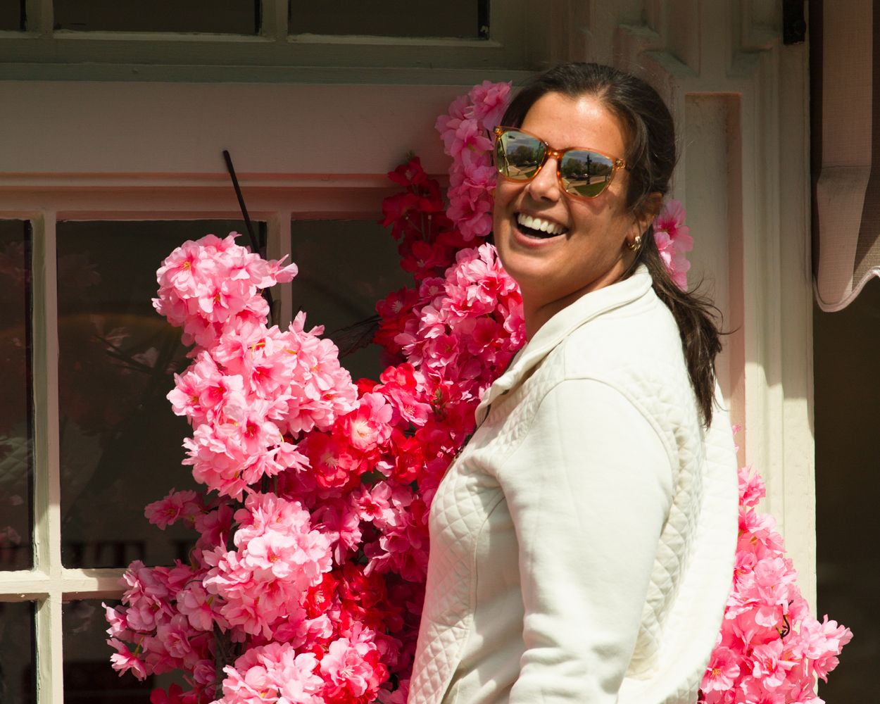 Laughing woman in white jacket posing beside vibrant pink cherry blossom floral display.