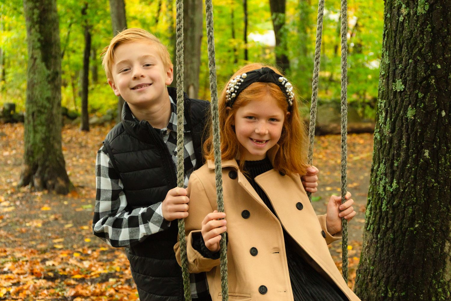 Smiling boy in a black vest and a red-haired girl in a tan coat hold ropes on a swing among autumn trees in a forest.