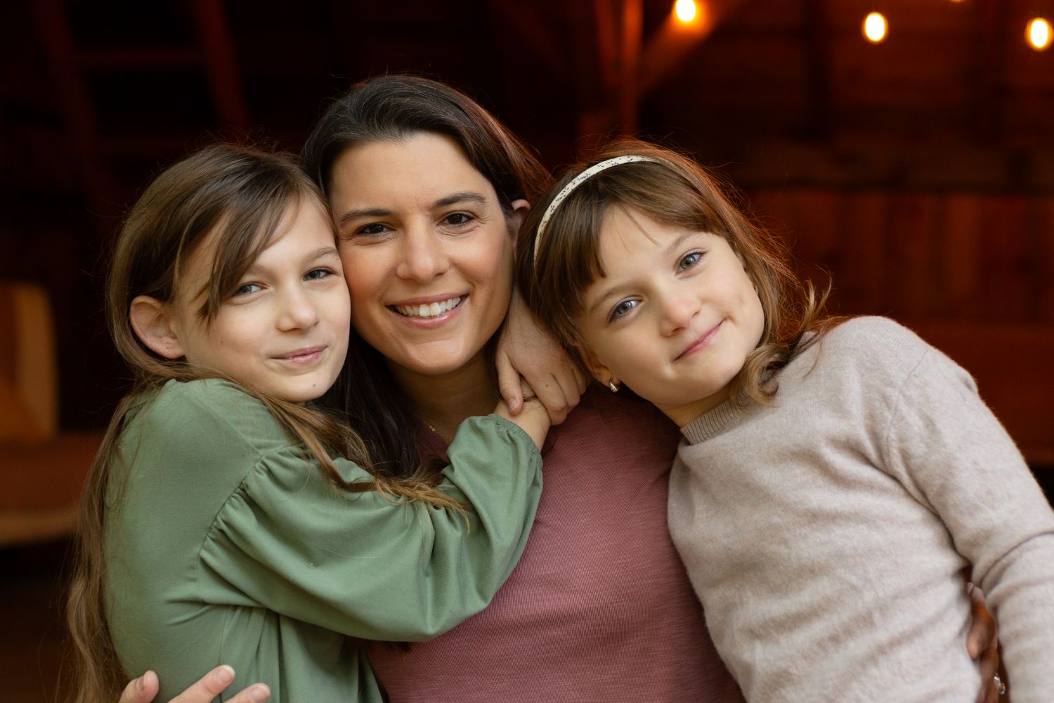 Smiling mother hugging two young daughters close in a cozy barn setting with warm string lights glowing in the background.