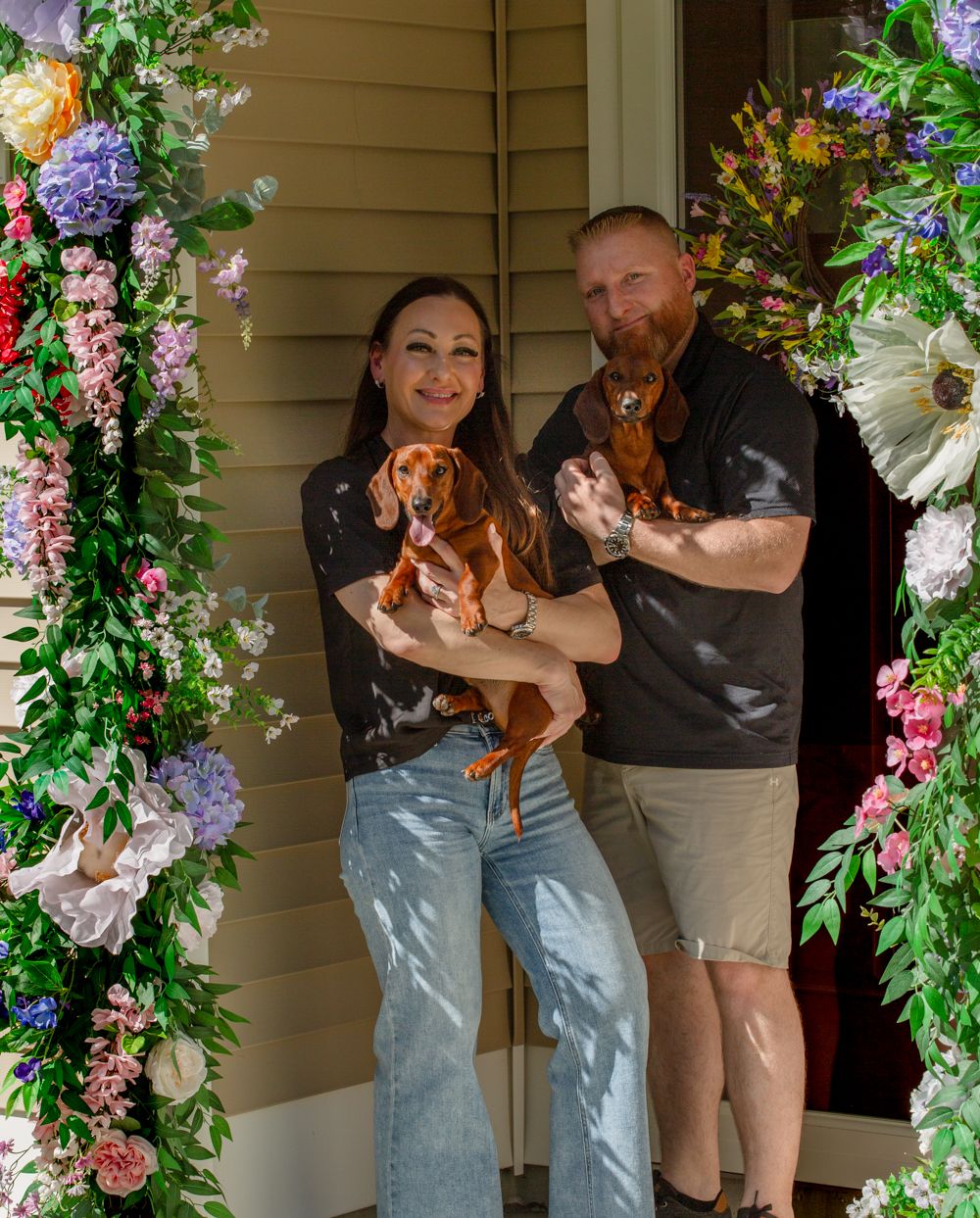Couple posing with two dachshunds framed by a vibrant floral arch at their front door.