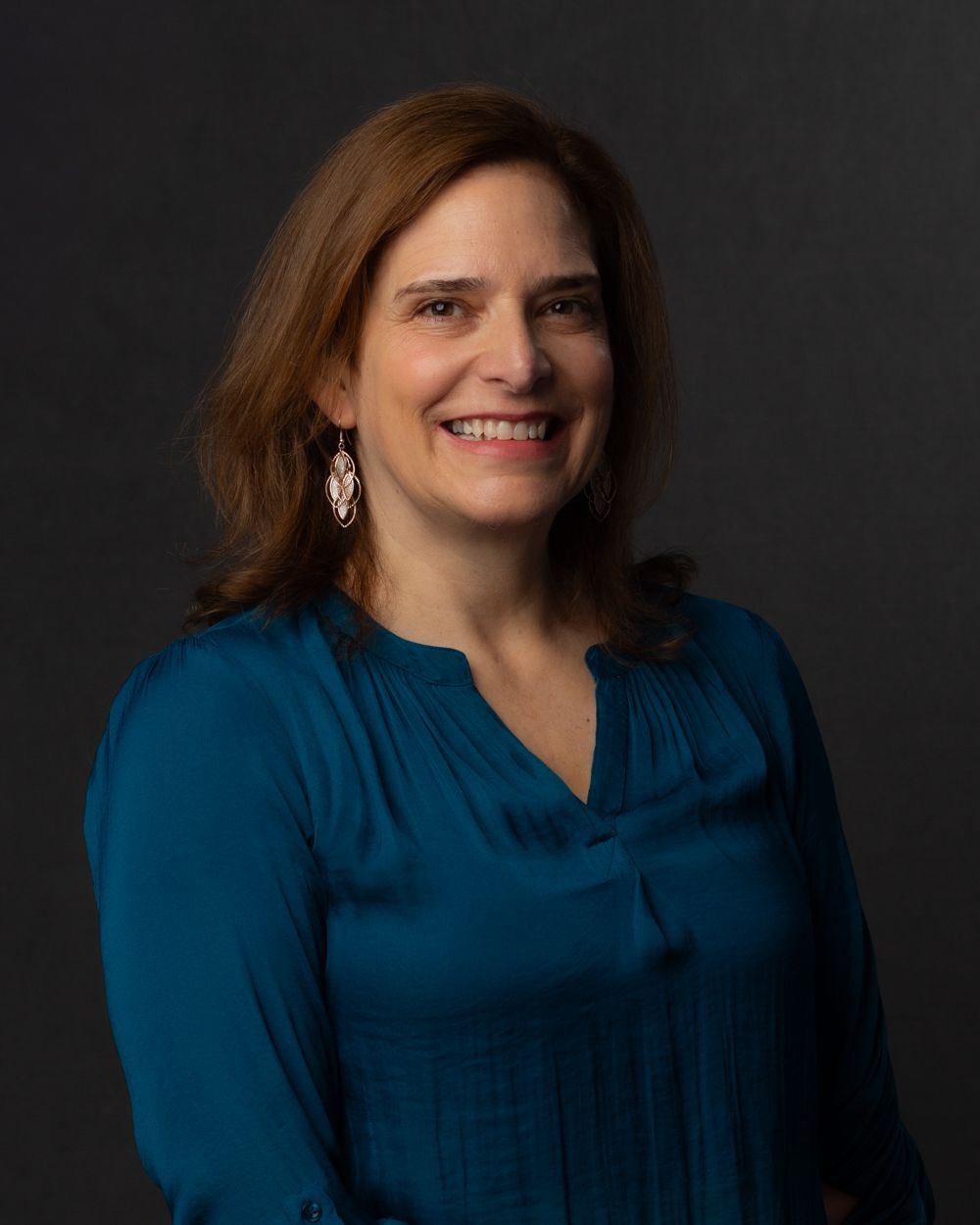 Professional woman in teal blouse with drop earrings smiling for headshot on dark background.