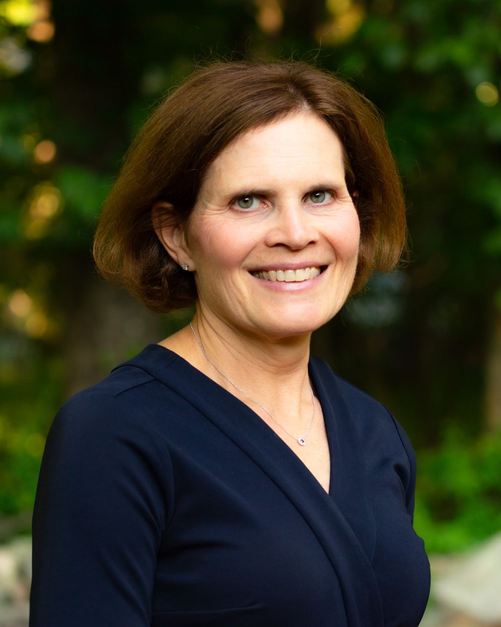 Professional headshot of smiling woman in navy blouse with green bokeh background.