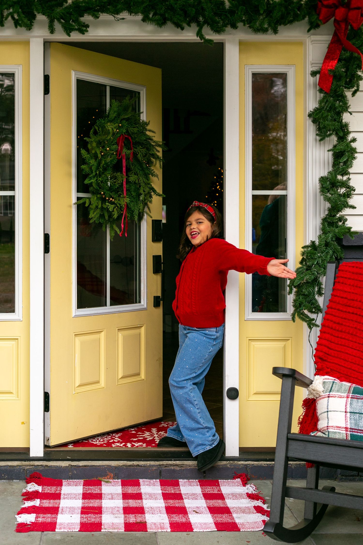 Woman in red sweater and jeans posing in Christmas-decorated yellow front door.