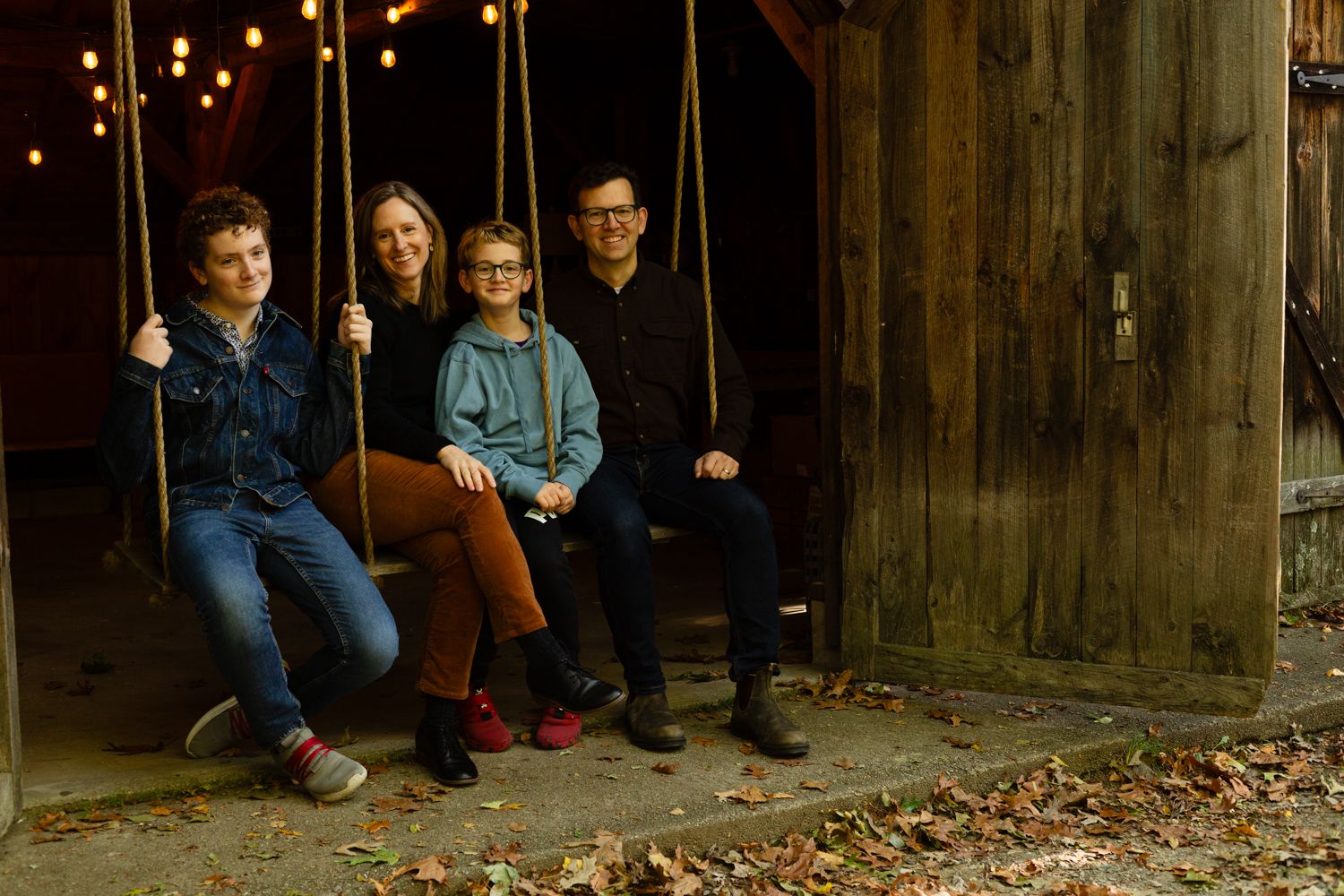 Family of four sits together on a rustic wooden swing beside a weathered barn, with warm string lights glowing in the background.
