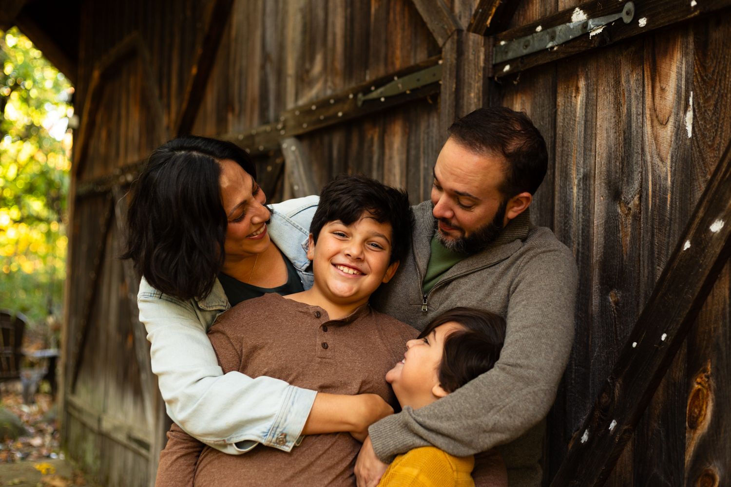 Happy family of four hugging closely in front of a rustic weathered wooden barn door on a sunny autumn day.