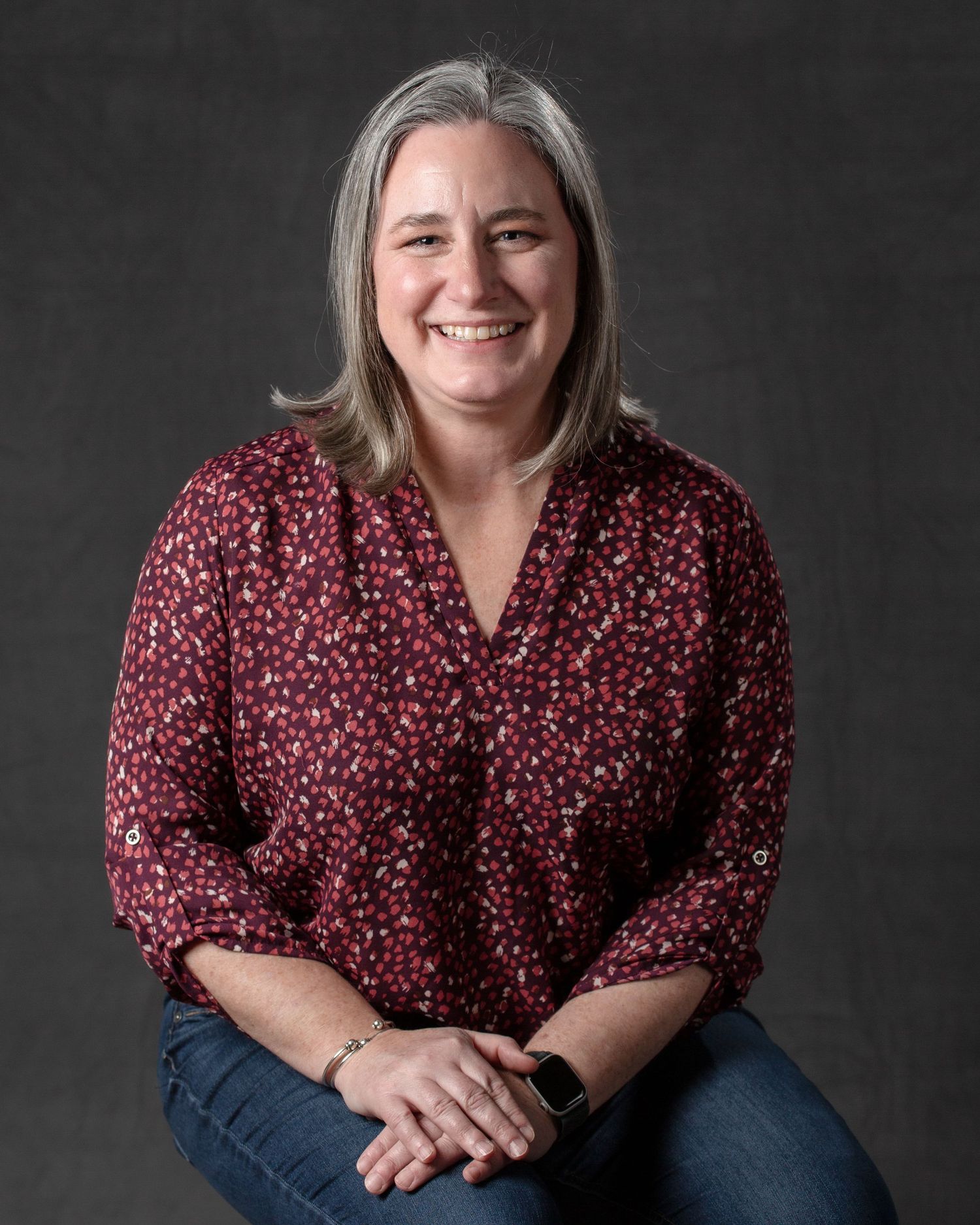 A smiling woman with gray-streaked hair wearing a red floral blouse poses for a professional photo.