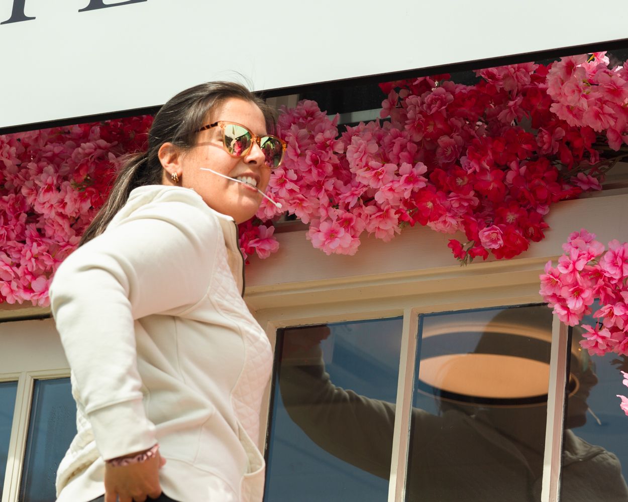 Smiling woman in white jacket and sunglasses standing in front of vibrant pink floral store display.