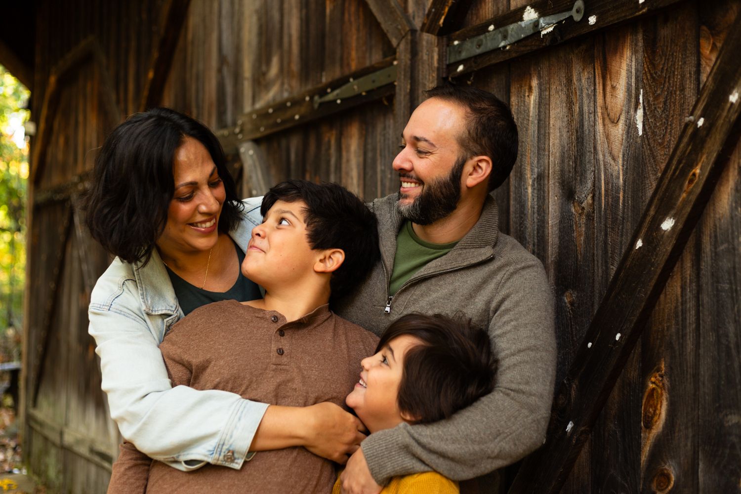 Smiling mother, father and two young sons embrace in a warm family portrait in front of a rustic weathered wooden barn.