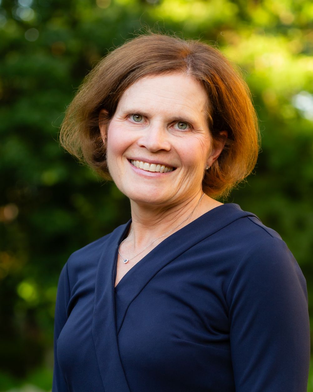 Smiling woman with short auburn hair wearing navy blue top outdoors with green foliage.