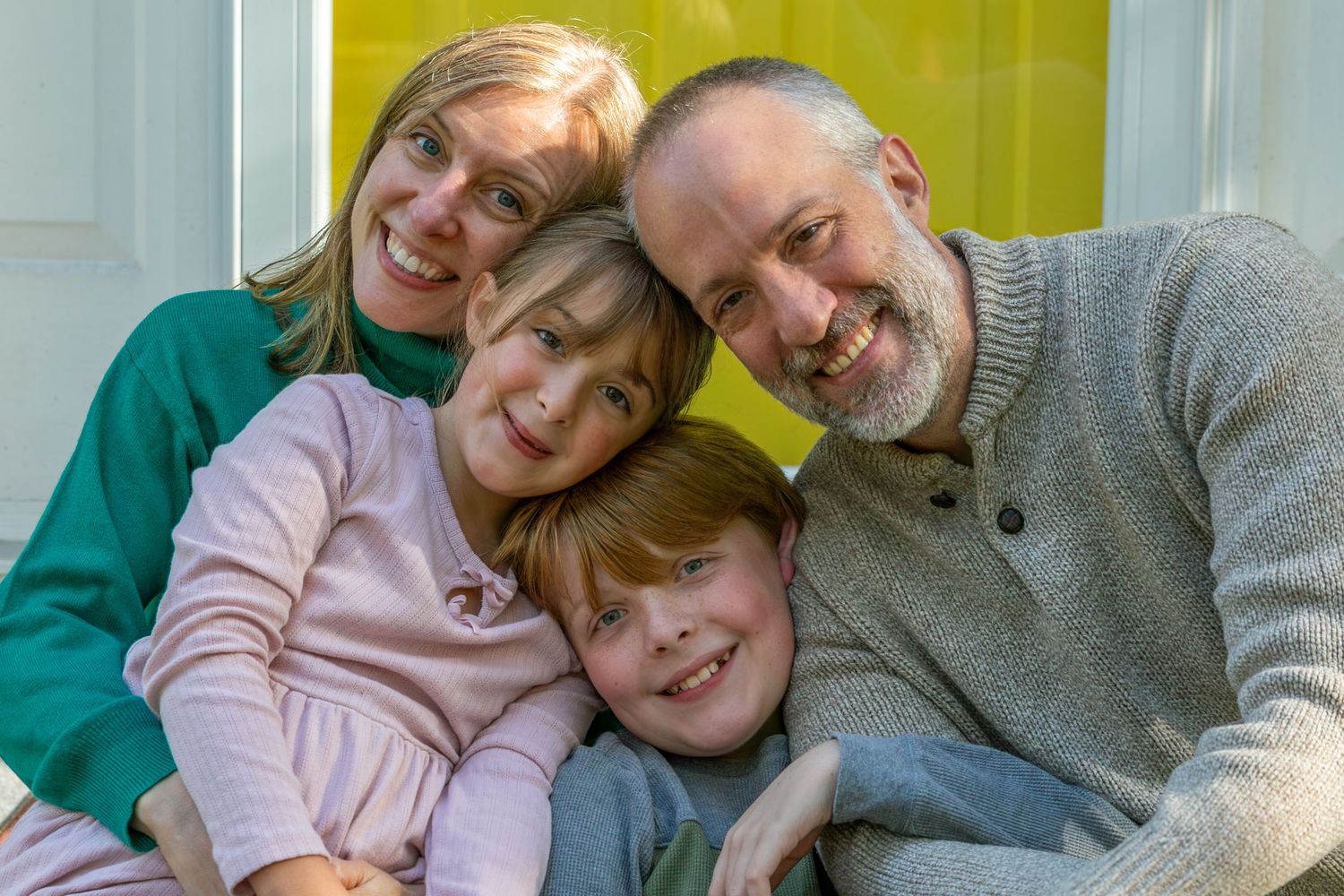 Happy family of four huddled together smiling in front of yellow door.