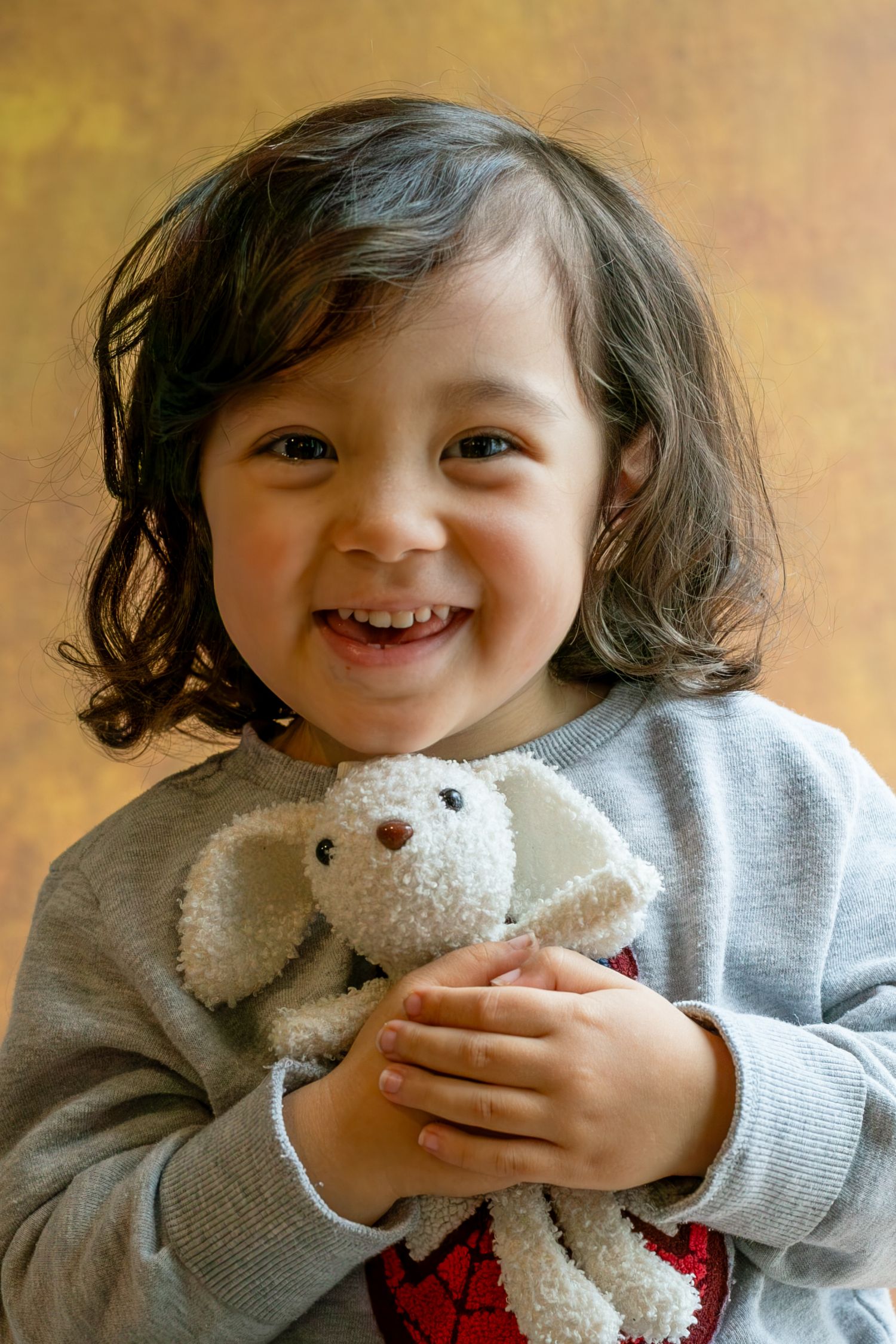 Happy toddler with dark curly hair smiling while holding a white stuffed bunny toy.
