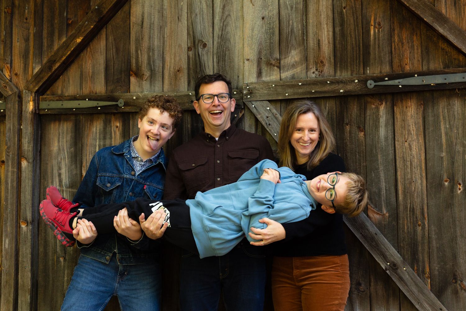 A smiling family of four poses in front of a rustic wooden barn door while holding a laughing child sideways in their arms.