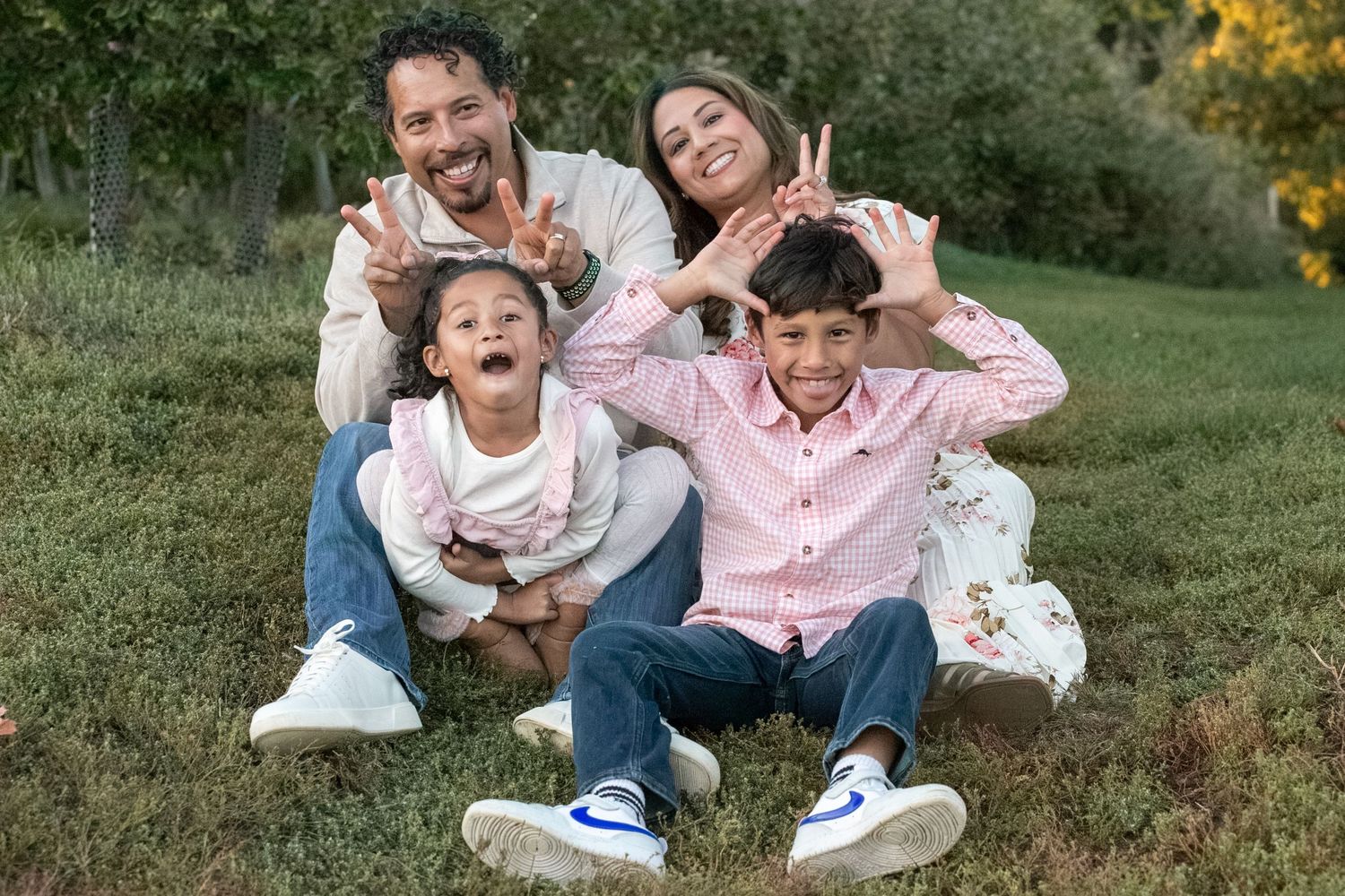 Happy family of four sitting on grass making funny faces and gestures outdoors in a park.