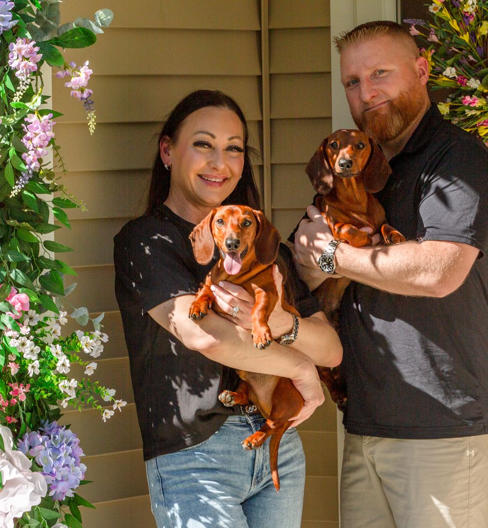 Happy couple holding two adorable dachshund dogs surrounded by colorful floral decorations.