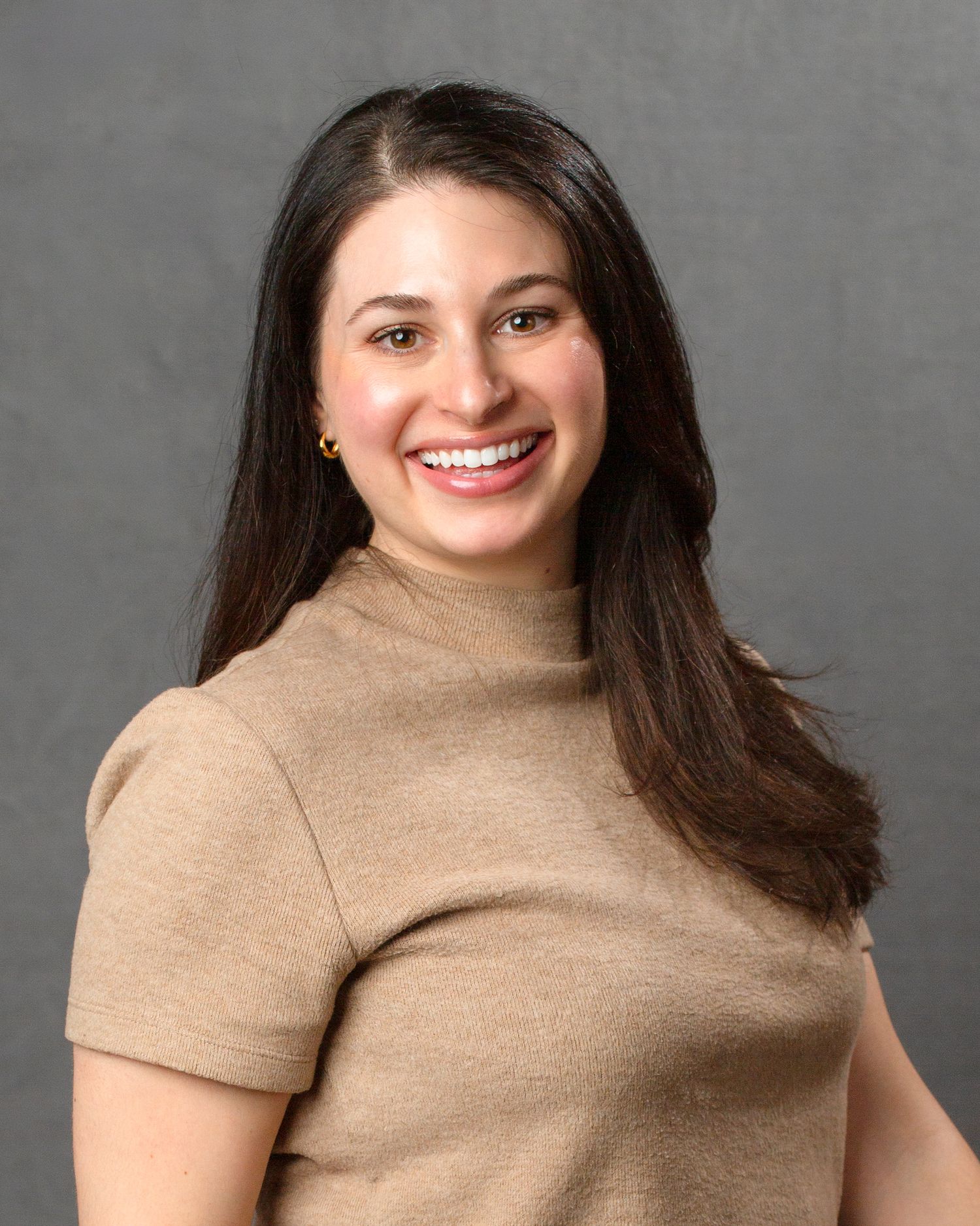Young woman in beige turtleneck top smiling for professional headshot on gray background.