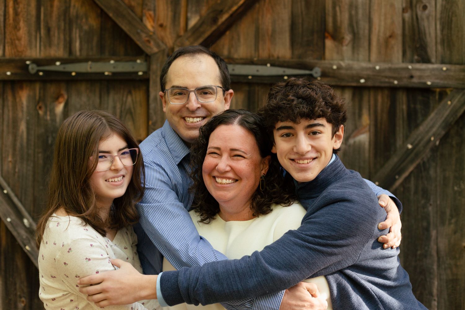 A smiling family of four hugging closely in front of a rustic weathered wooden barn door during an outdoor portrait session.