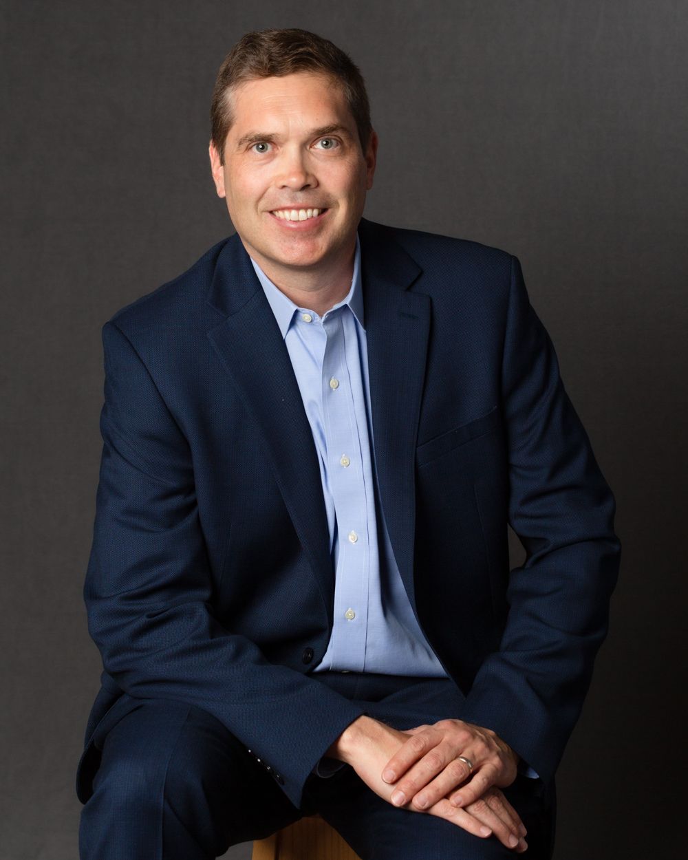 Professional headshot of a smiling man in a navy blue blazer and light blue shirt seated against a gray background.