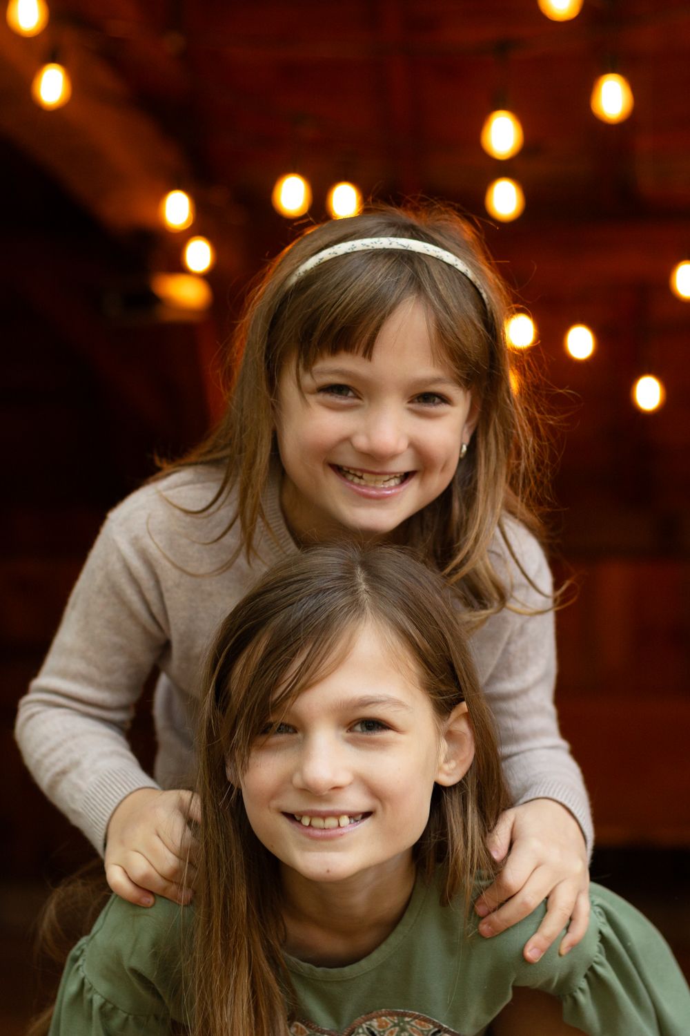 Two smiling sisters pose together, one standing behind the other with hands on her shoulders, beneath warm glowing string lights.
