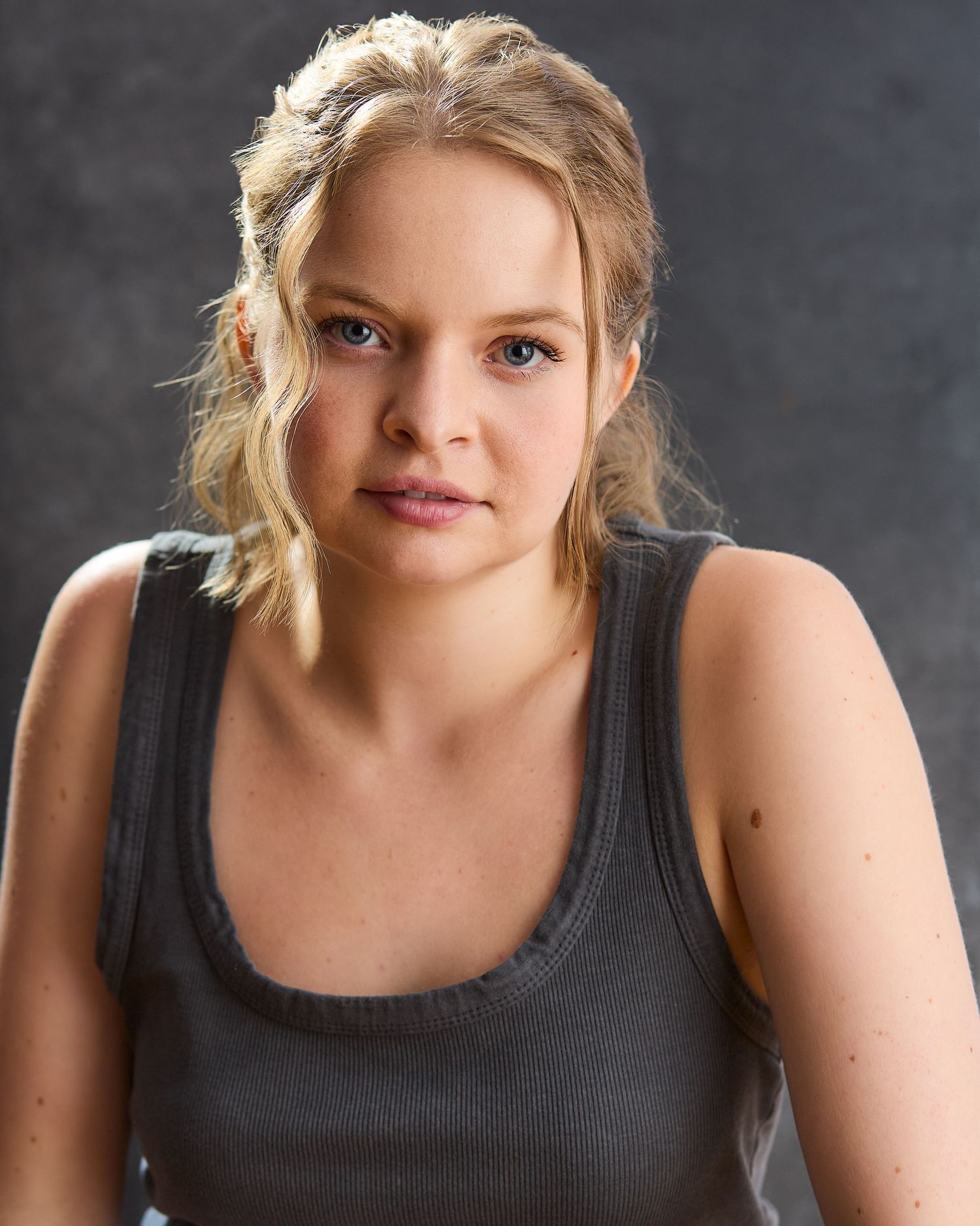 A young blonde woman in a gray tank top poses for a acting headshot against a neutral background.