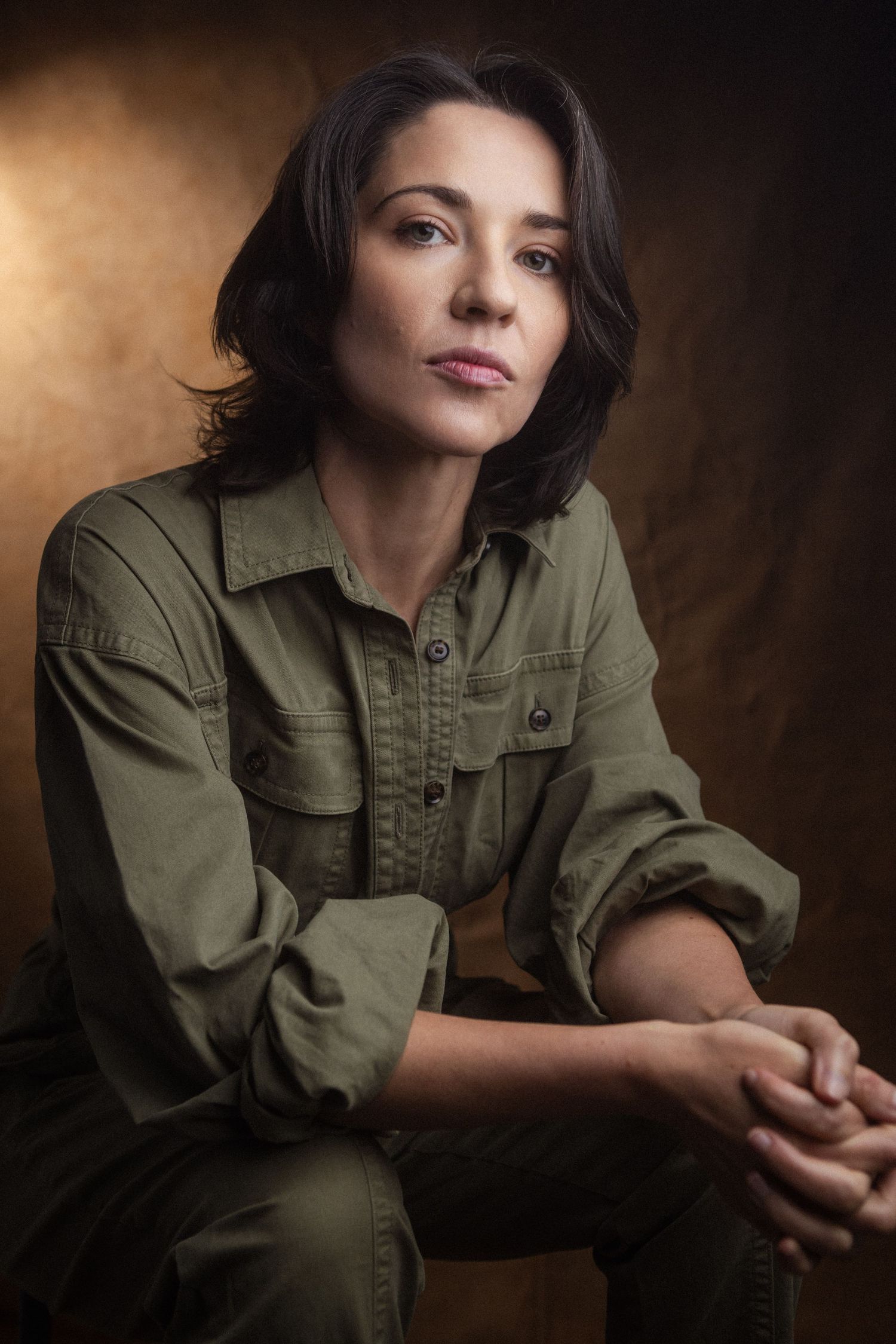 Woman in olive green utility shirt seated confidently against warm brown studio background.