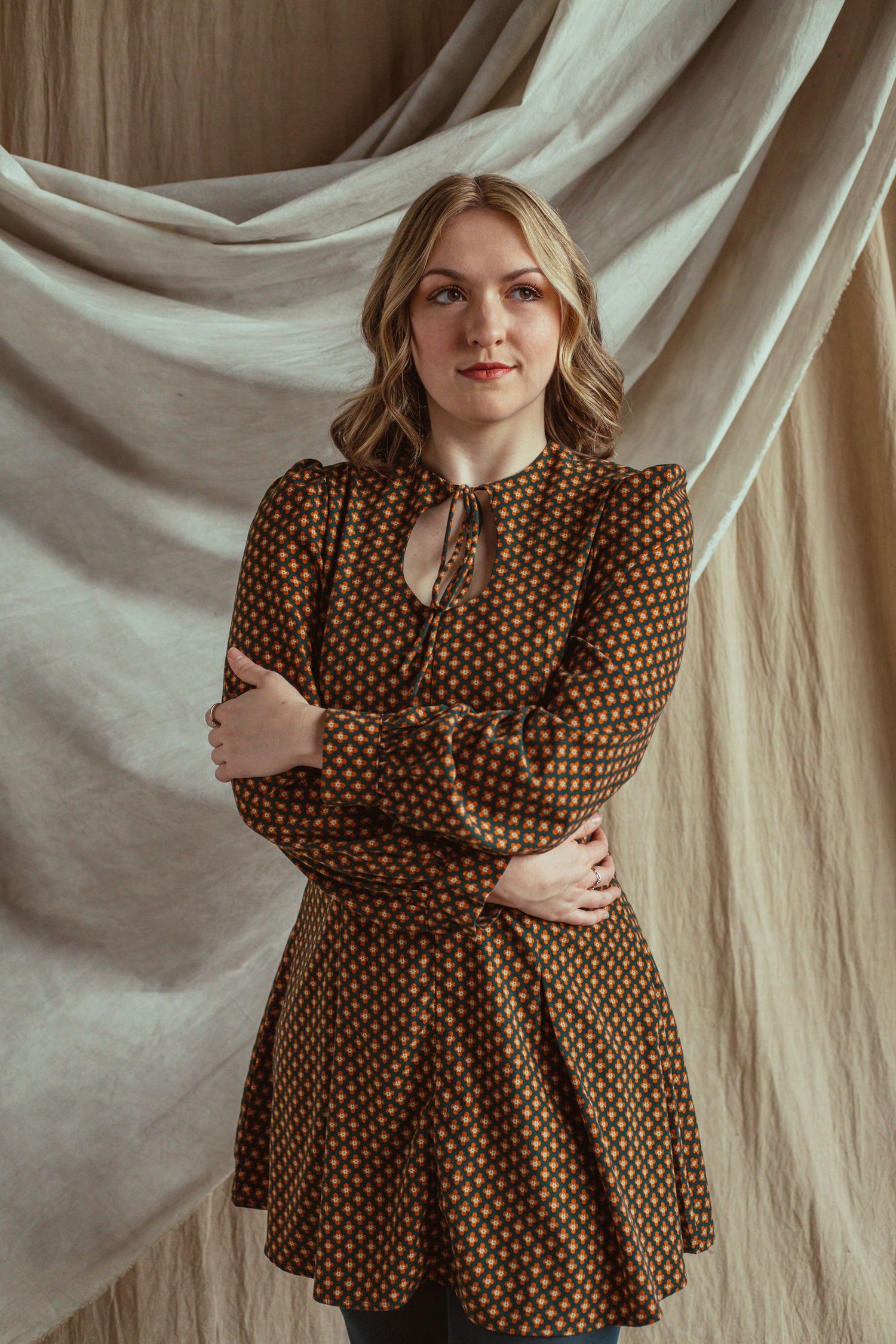 Blonde woman in brown polka dot dress with puffed sleeves poses with arms crossed before draped fabric.