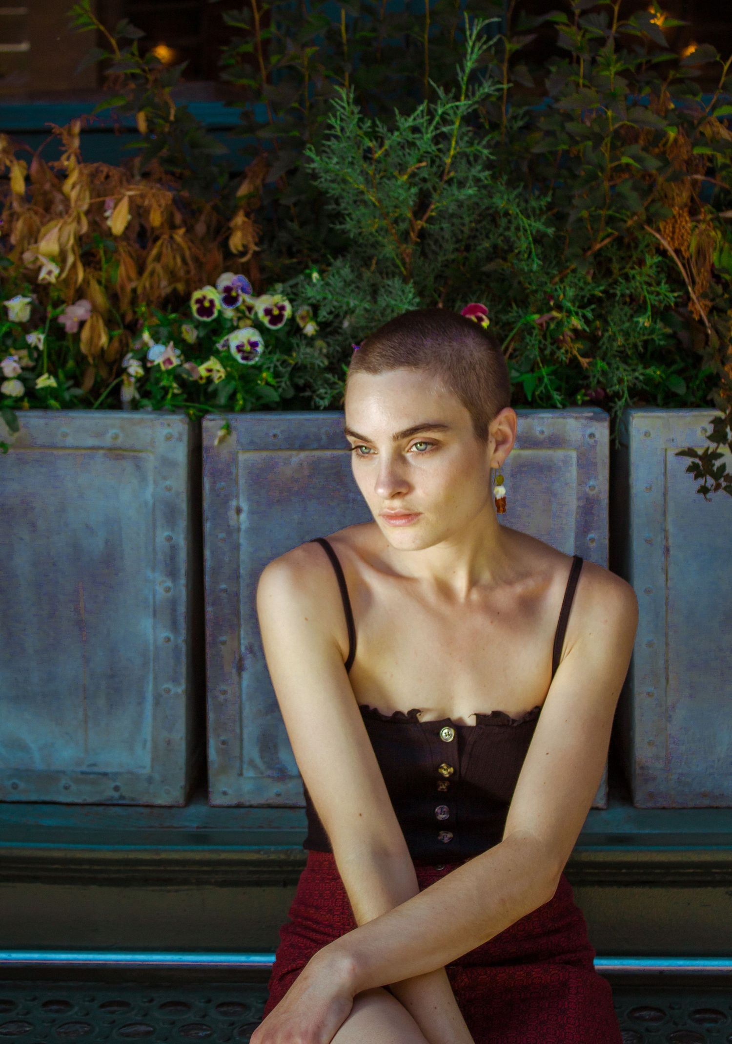 A woman with a shaved head in a black camisole sits on a bench surrounded by flowers.