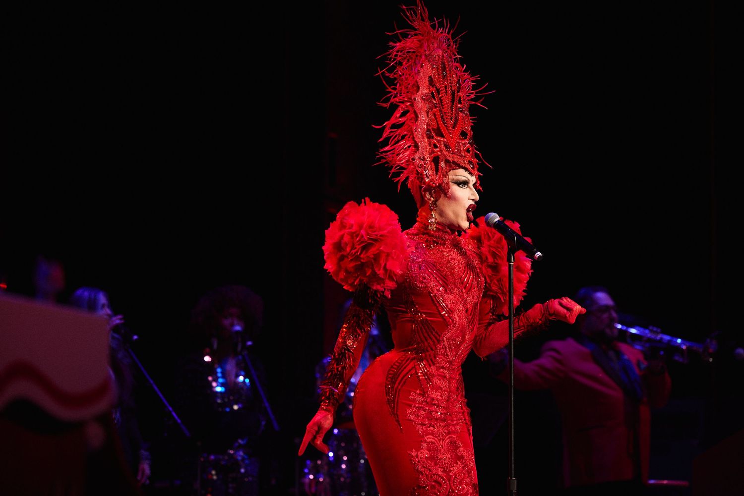 Drag Queen Sasha Velour in an elaborate red feathered costume and headdress sings into a microphone on stage.