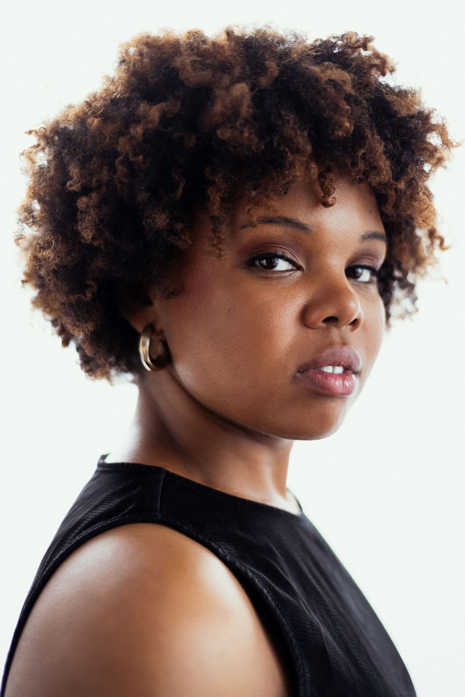 Young Black woman with natural curly afro hair wearing a black sleeveless top and gold hoop earrings.
