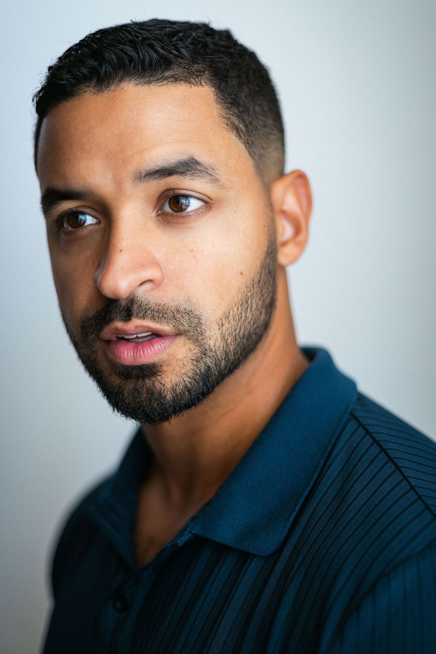 Close-up portrait of a young man with short dark hair and beard wearing a teal polo shirt against a light background.