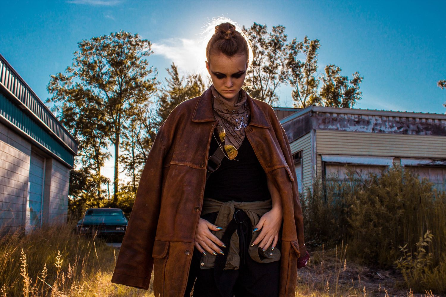 Woman in a brown leather jacket stands outdoors at golden hour near an abandoned building.
