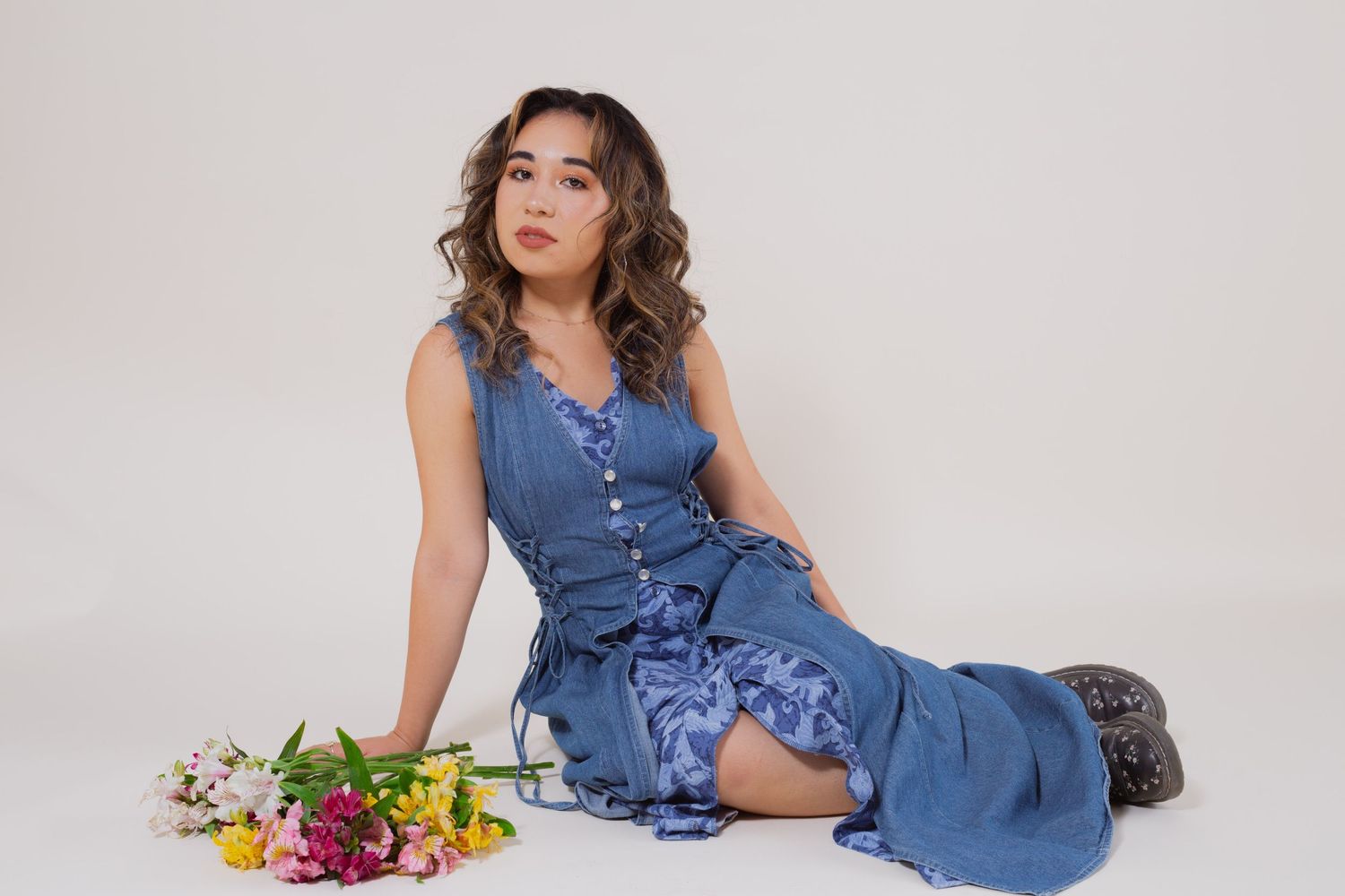 Young woman in a blue dress sitting on floor with colorful flowers beside her in a white studio.