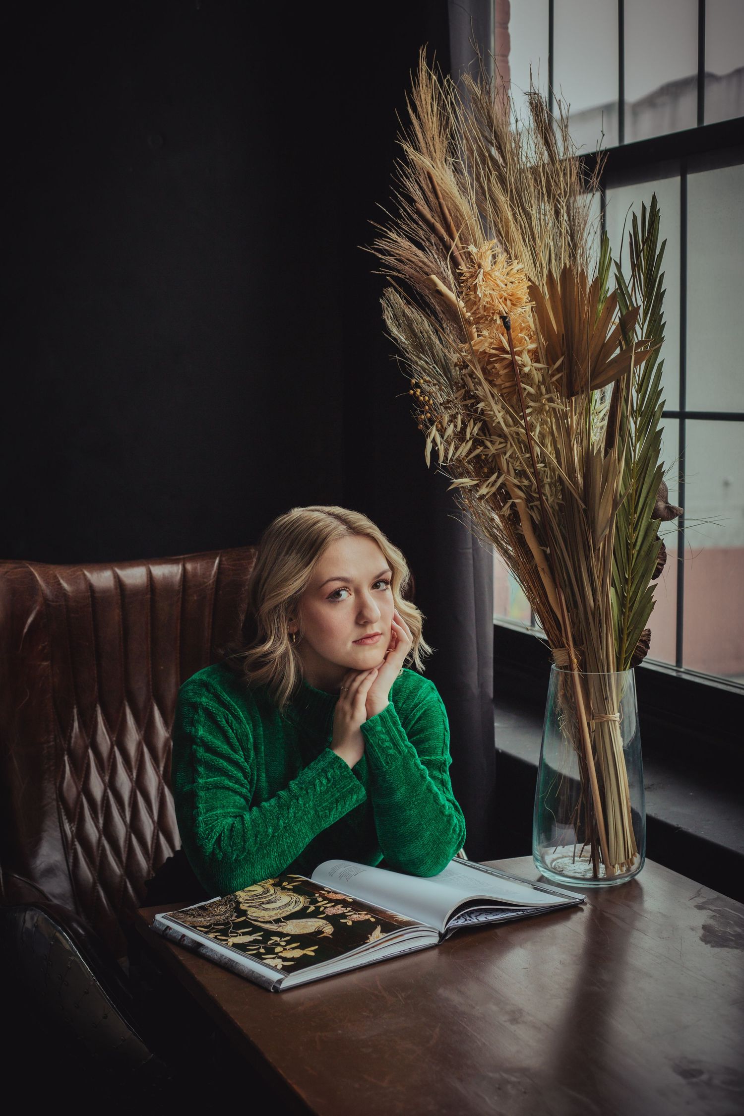 Blonde woman in green knit sweater sits at table with open book and dried floral arrangement.