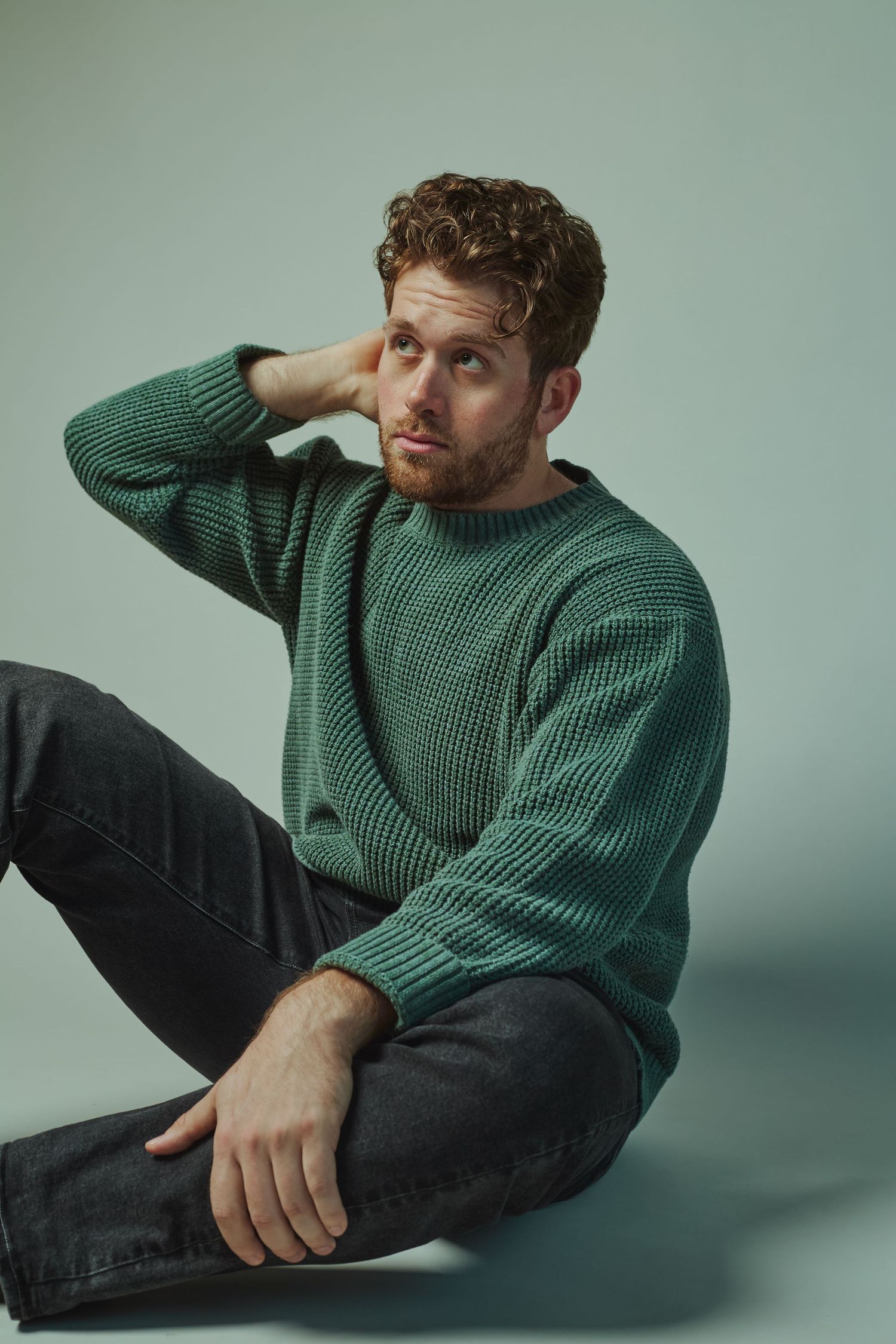 Redheaded man in a green knit sweater sits casually against a light green studio background.