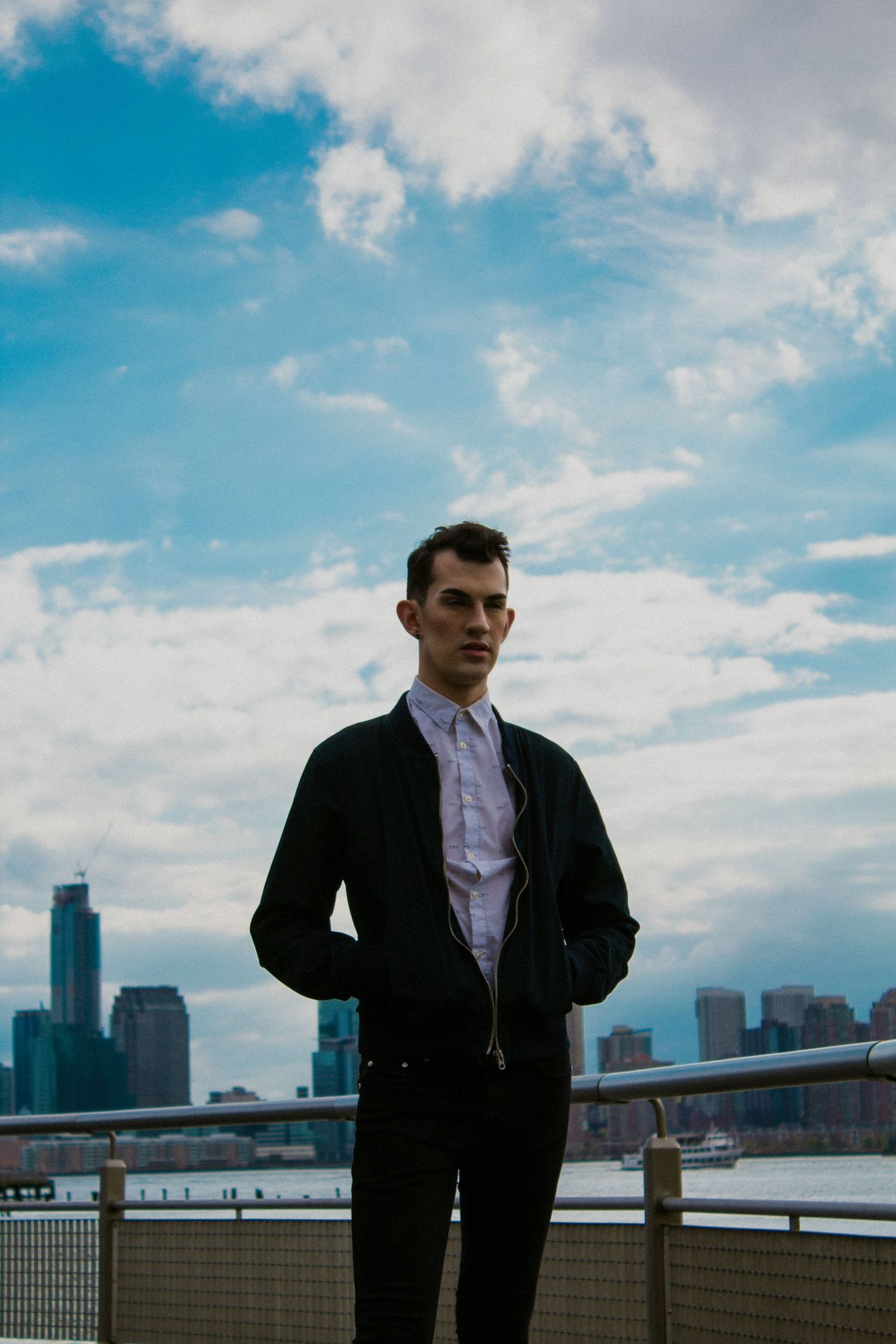 Person in black jacket and white shirt stands before a city skyline under cloudy blue sky.