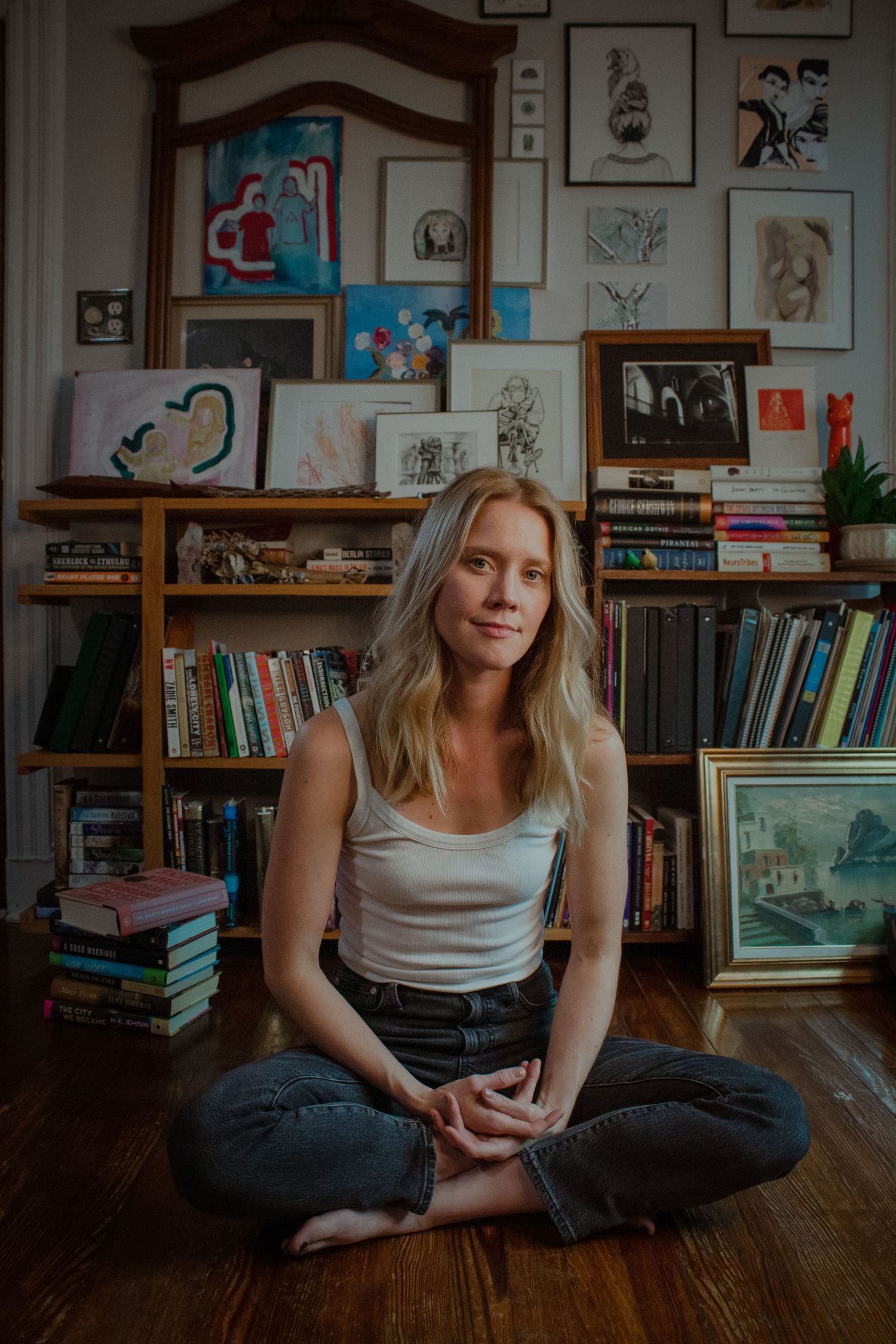 Blonde woman sits cross-legged on a wood floor in front of bookshelves filled with art and books.
