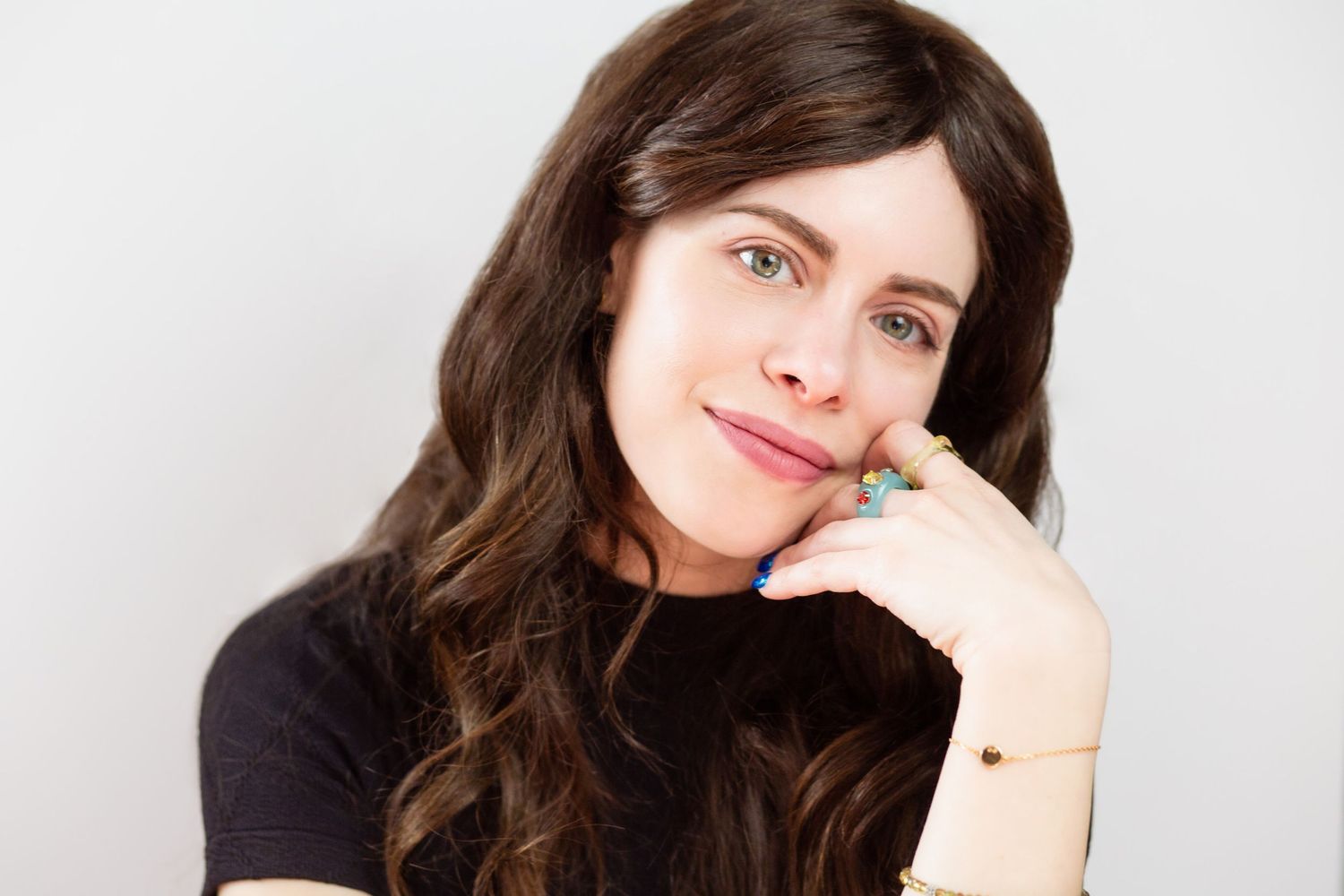 Brunette woman with wavy hair resting chin on hands, smiling softly against a white background.