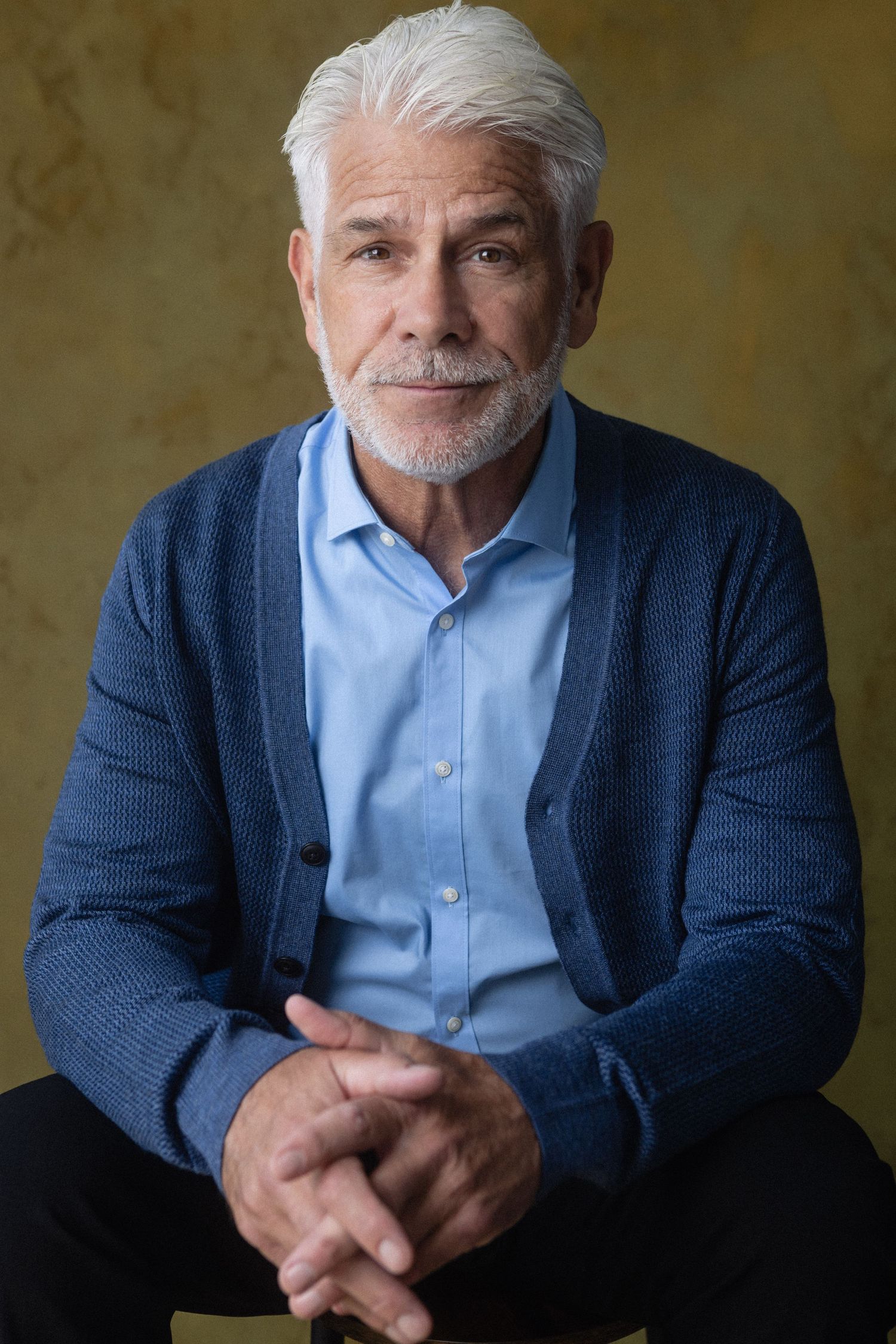 Confident senior man with white hair and beard wearing a blue cardigan over a light blue shirt, sitting with hands clasped.