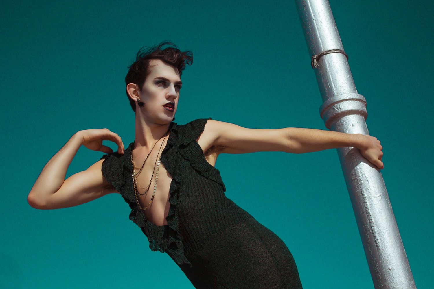 Model in dark green dress leans against a metal pole against a teal background.