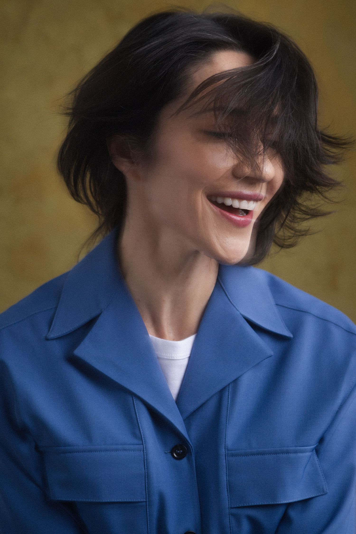 Laughing woman with short dark hair wearing a blue jacket over a white shirt against a yellow background.